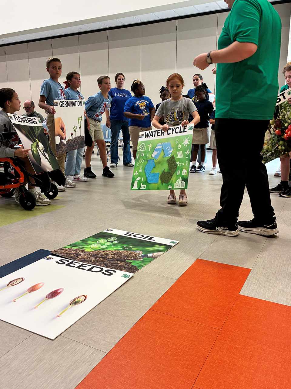 A group of children are gathered around another child who is holding up a sign with a picture of a water cycle.